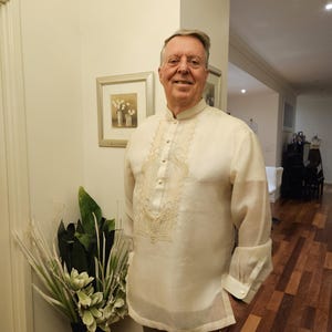 May include: A cream-colored long-sleeved barong tagalog shirt featuring a mandarin collar, a button-down front placket, and intricate tonal embroidery patterns along the chest area. The garment is made of a sheer, lightweight fabric.