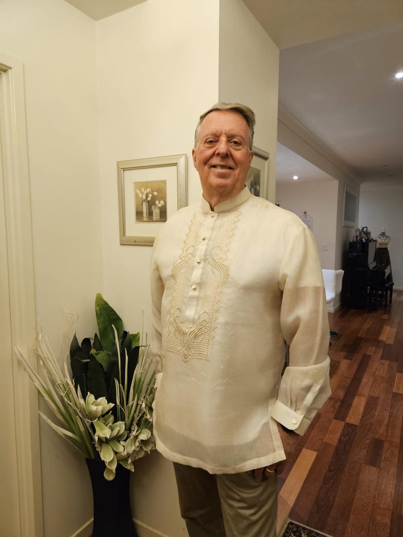 May include: A cream-colored long-sleeved barong tagalog shirt featuring a mandarin collar, a button-down front placket, and intricate tonal embroidery patterns along the chest area. The garment is made of a sheer, lightweight fabric.