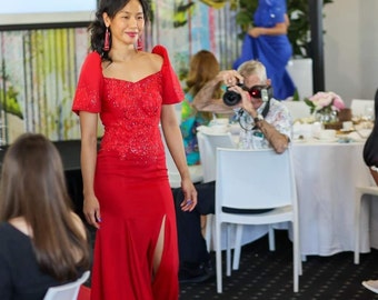 May include: A woman in a red, floor-length gown with a high slit, walking on a red carpet. The dress features a detailed bodice and short sleeves. The background includes a blurred event setting with tables and people.