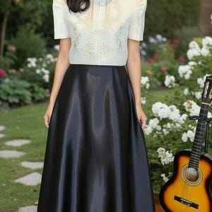 May include: A woman wearing a cream-colored top with intricate embroidery and a high collar, paired with a long, black satin skirt. A guitar rests nearby, suggesting a musical theme. The outfit is suitable for formal events.
