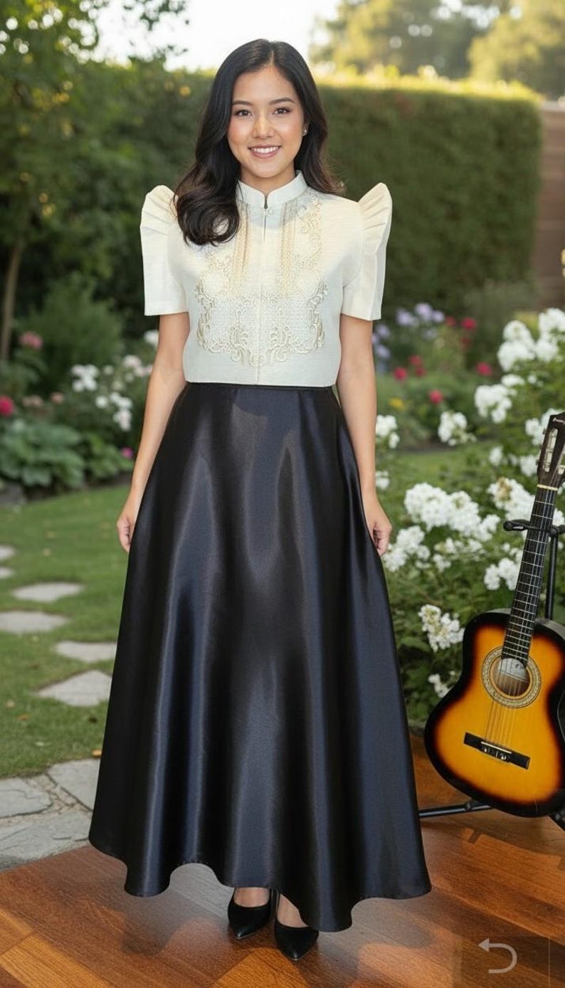 May include: A woman wearing a cream-colored top with intricate embroidery and a high collar, paired with a long, black satin skirt. A guitar rests nearby, suggesting a musical theme. The outfit is suitable for formal events.