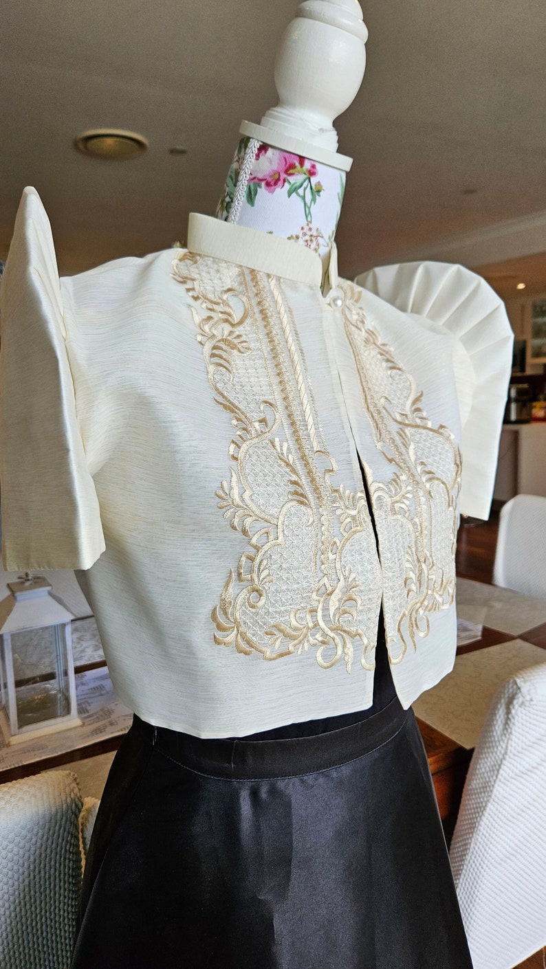 May include: Ivory-colored formal top with intricate gold embroidery. The top features short sleeves and a high collar. It is paired with a black skirt, creating a classic and elegant ensemble. The outfit is displayed on a mannequin.