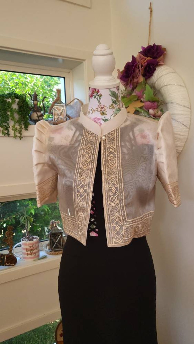 May include: A cream-colored bolero jacket with sheer fabric, featuring structured puff sleeves and intricate gold-toned embroidery along the front panels and cuffs. The jacket has a high collar, a single pearl-like button closure, and is displayed on a mannequin.