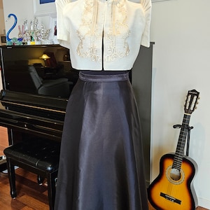 May include: A cream-colored, short-sleeved top with intricate gold embroidery and a high collar, paired with a long, black satin skirt. The outfit is displayed on a mannequin, showcasing a traditional formal ensemble.