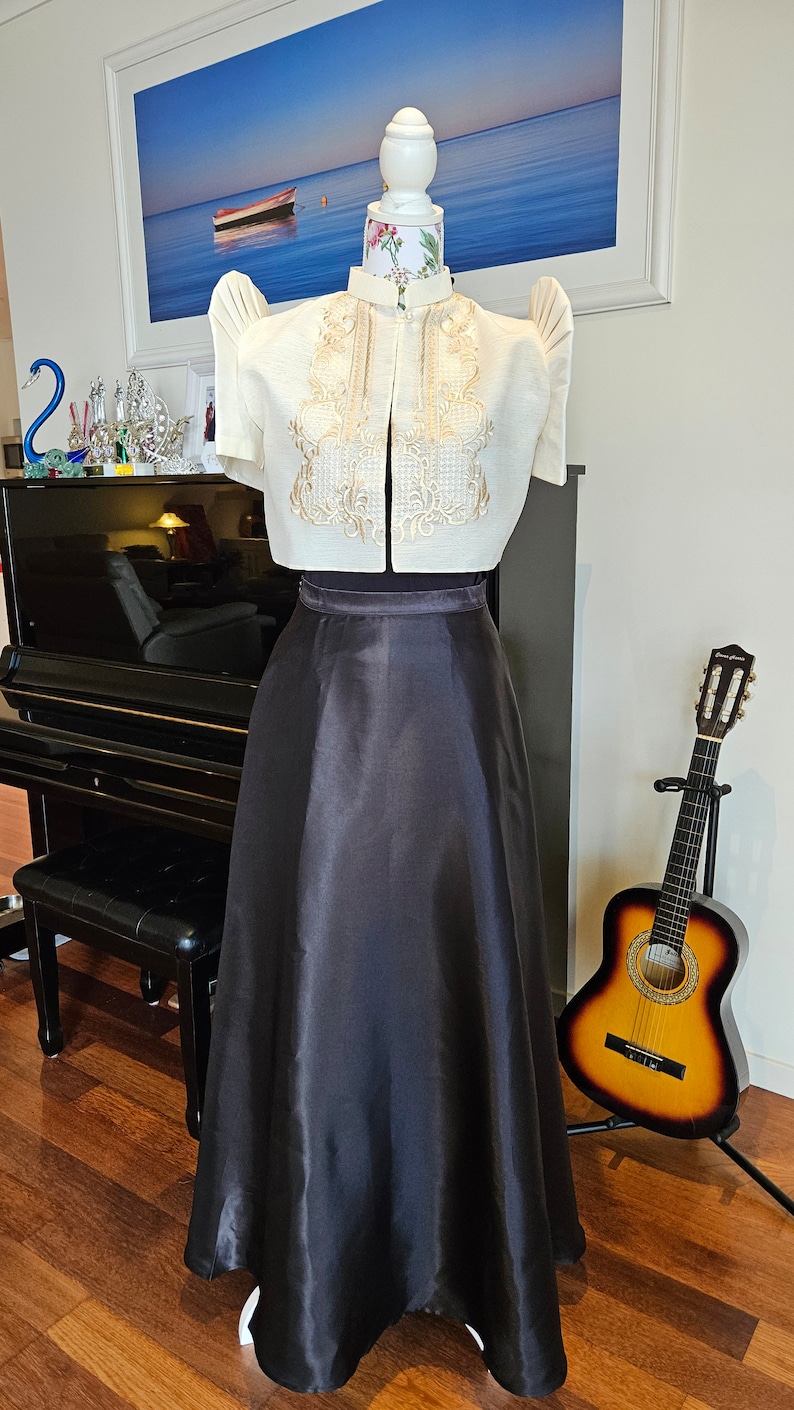 May include: A cream-colored, short-sleeved top with intricate gold embroidery and a high collar, paired with a long, black satin skirt. The outfit is displayed on a mannequin, showcasing a traditional formal ensemble.