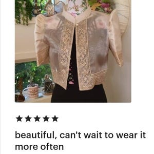 May include: A beige bolero jacket featuring sheer fabric, structured puff sleeves, and intricate cream-colored embroidery along the front panels and sleeve cuffs. The garment has a mandarin collar and a single pearl-like button closure at the neck.