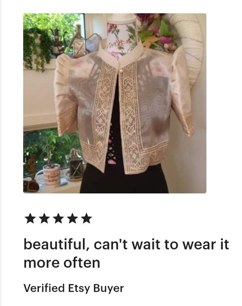 May include: A beige bolero jacket featuring sheer fabric, structured puff sleeves, and intricate cream-colored embroidery along the front panels and sleeve cuffs. The garment has a mandarin collar and a single pearl-like button closure at the neck.