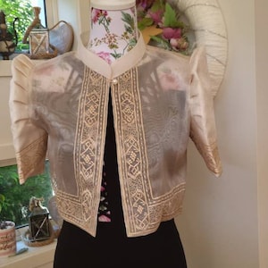 May include: A sheer, cream-colored bolero jacket featuring short, puffed sleeves and intricate, light-colored embroidery along the front panels and sleeve cuffs. The garment has a high collar and a single pearl-like button closure at the neckline.