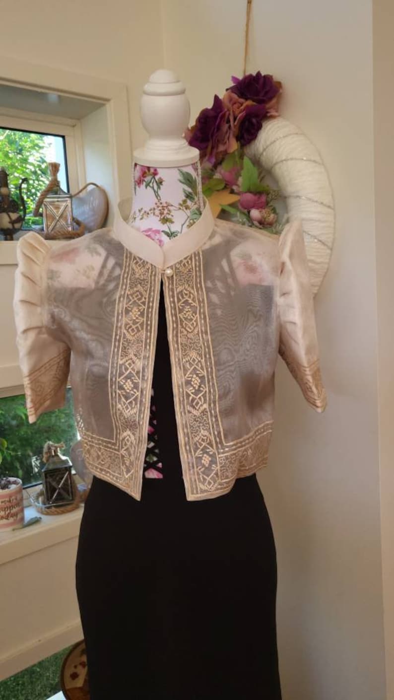 May include: A sheer, cream-colored bolero jacket featuring short, puffed sleeves and intricate, light-colored embroidery along the front panels and sleeve cuffs. The garment has a high collar and a single pearl-like button closure at the neckline.