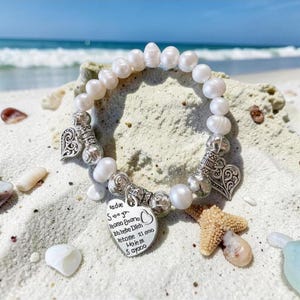 May include: A pearl bracelet with silver heart charms and beads, resting on a sandy beach with a starfish and seashells. The bracelet has a heart-shaped charm with engraved text.