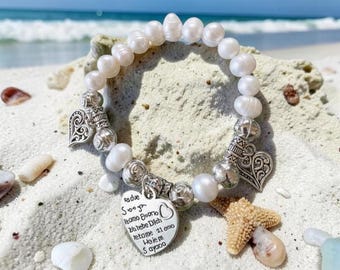 May include: A pearl bracelet with silver heart charms and beads, resting on a sandy beach with a starfish and seashells. The bracelet has a heart-shaped charm with engraved text.