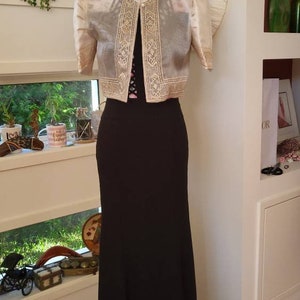 May include: A floor-length black dress paired with a cropped, sheer, cream-colored bolero jacket. The jacket features short, puffed sleeves and intricate gold-toned embroidery along the front panels, collar, and sleeve cuffs, secured by a single button.