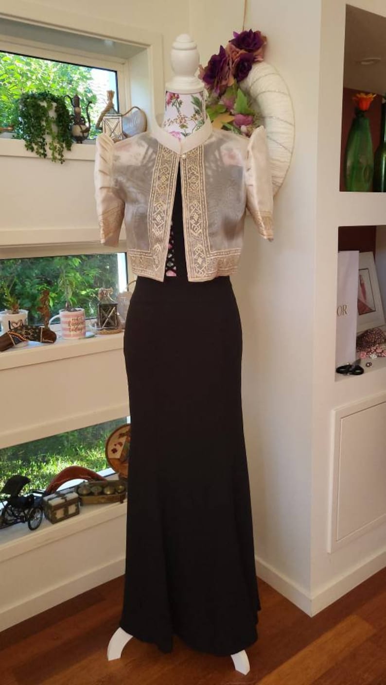May include: A floor-length black dress paired with a cropped, sheer, cream-colored bolero jacket. The jacket features short, puffed sleeves and intricate gold-toned embroidery along the front panels, collar, and sleeve cuffs, secured by a single button.