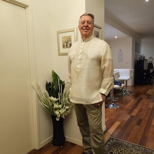 May include: A cream-colored long-sleeved barong tagalog shirt featuring a mandarin collar, a button-down front placket, and intricate tonal embroidery on the chest, paired with light brown trousers and dark brown dress shoes.