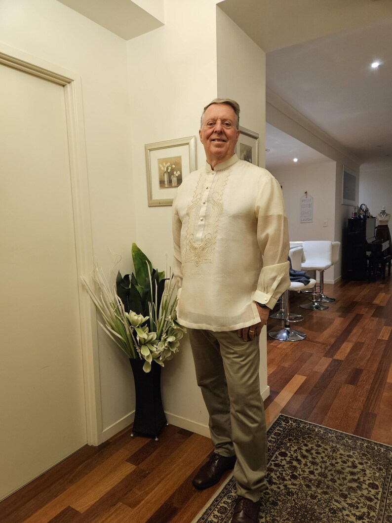 May include: A cream-colored long-sleeved barong tagalog shirt featuring a mandarin collar, a button-down front placket, and intricate tonal embroidery on the chest, paired with light brown trousers and dark brown dress shoes.
