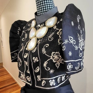 以下が含まれることがあります： 白い花の刺繍とパフスリーブが付いた黒いブラウスがマネキンに展示されています。 ブラウスはハイカラーと、大きくて白い楕円形の石が付いた装飾的なネックレスが特徴です。 マネキンの首は白黒のチェック柄の生地で覆われています。