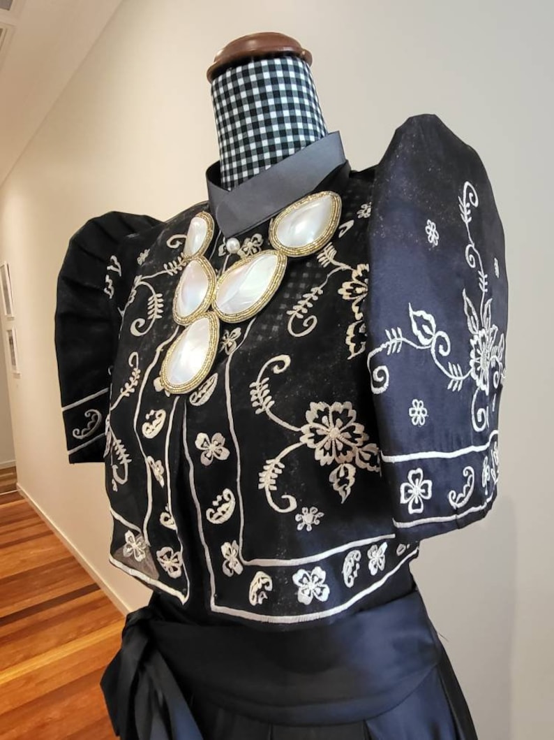 以下が含まれることがあります： 白い花の刺繍とパフスリーブが付いた黒いブラウスがマネキンに展示されています。 ブラウスはハイカラーと、大きくて白い楕円形の石が付いた装飾的なネックレスが特徴です。 マネキンの首は白黒のチェック柄の生地で覆われています。