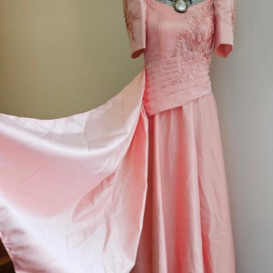 May include: A light pink formal gown with short sleeves and a flowing skirt. The bodice features intricate beadwork and a pleated waist. A large, pearlescent necklace adorns the mannequin's neck. The dress is displayed on a white mannequin.
