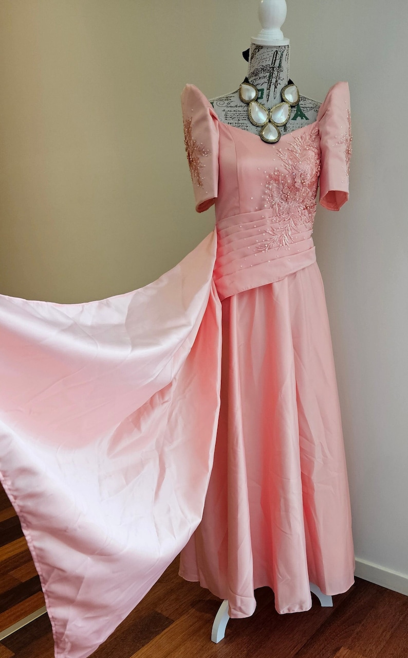 May include: A light pink formal gown with short sleeves and a flowing skirt. The bodice features intricate beadwork and a pleated waist. A large, pearlescent necklace adorns the mannequin's neck. The dress is displayed on a white mannequin.