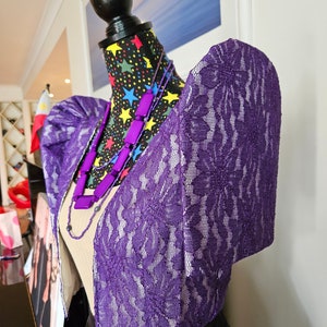 May include: A purple lace bolero jacket featuring structured butterfly sleeves, worn over a beige top. The garment is displayed on a mannequin with a black fabric cover patterned with colorful stars, accessorized with a purple beaded necklace.