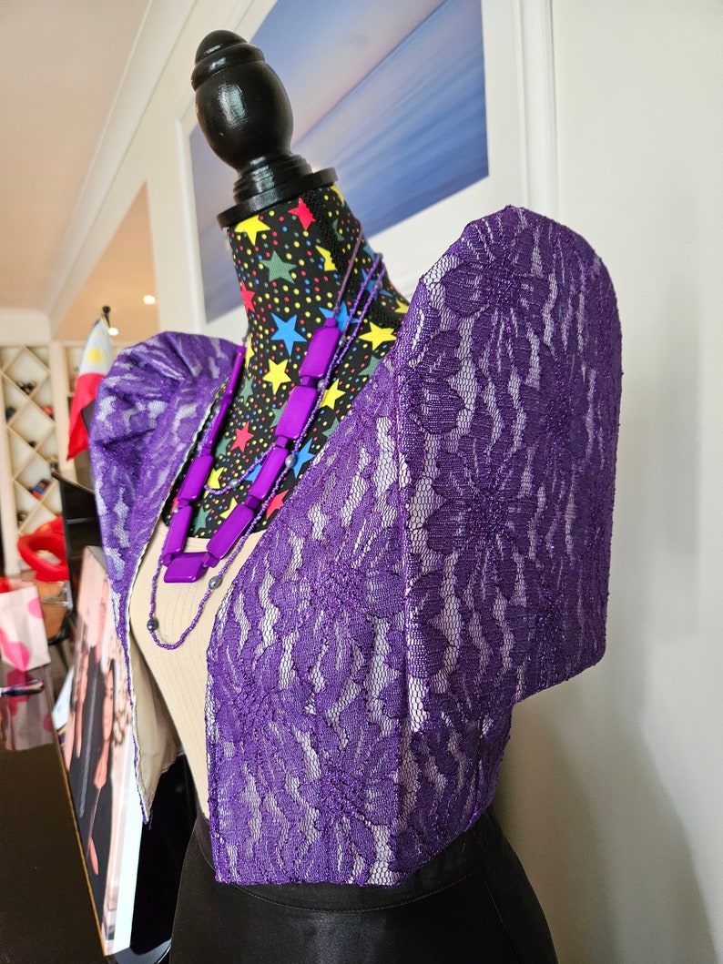 May include: A purple lace bolero jacket featuring structured butterfly sleeves, worn over a beige top. The garment is displayed on a mannequin with a black fabric cover patterned with colorful stars, accessorized with a purple beaded necklace.