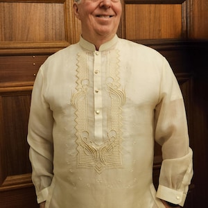 May include: A long-sleeved, off-white Barong Tagalog shirt featuring a mandarin collar, a button-down placket, and intricate cream-colored embroidery along the chest panel. The garment is crafted from a sheer, lightweight, and semi-transparent fabric.