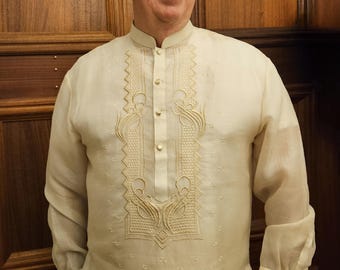 May include: A long-sleeved, off-white Barong Tagalog shirt featuring a mandarin collar, a button-down placket, and intricate cream-colored embroidery along the chest panel. The garment is crafted from a sheer, lightweight, and semi-transparent fabric.