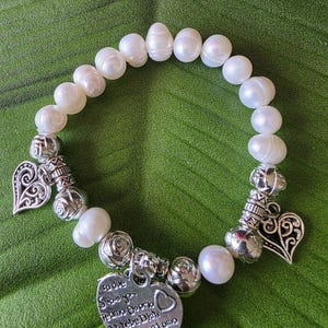 Peut inclure: Bracelet de perles blanches avec des breloques en forme de c&oelig;ur argent&eacute;es et des perles d&eacute;coratives. Le bracelet comporte un charm en forme de c&oelig;ur avec du texte grav&eacute;. Sur fond vert.