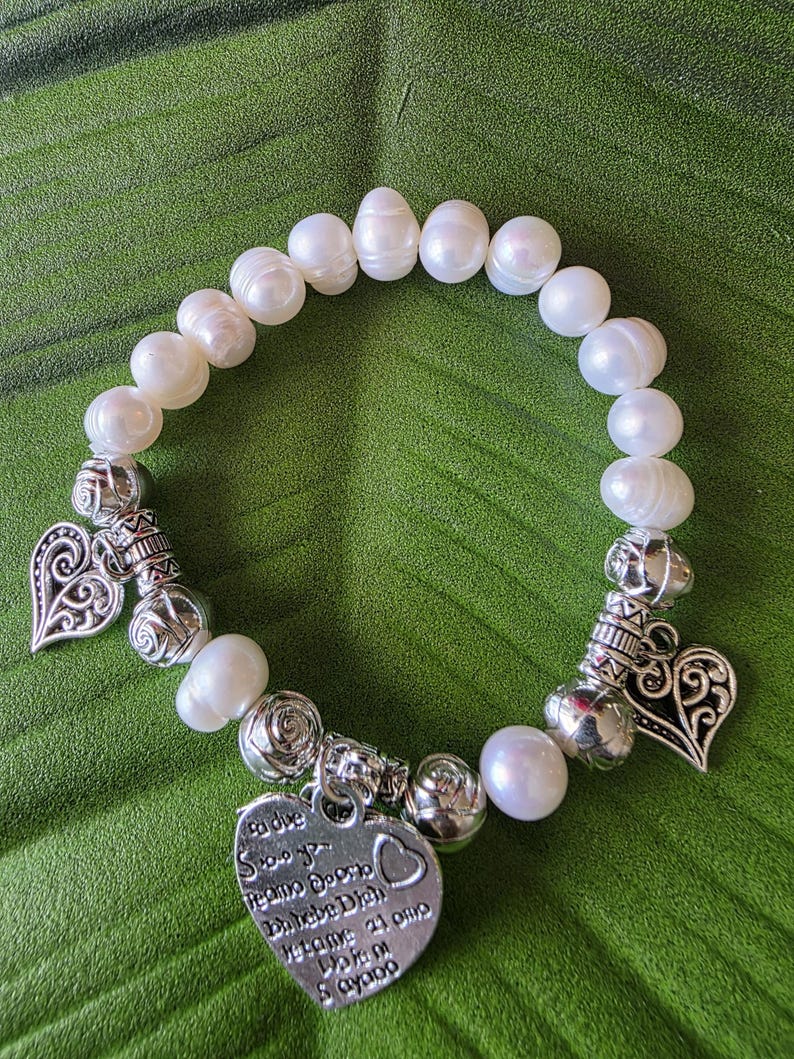 Peut inclure: Bracelet de perles blanches avec des breloques en forme de c&oelig;ur argent&eacute;es et des perles d&eacute;coratives. Le bracelet comporte un charm en forme de c&oelig;ur avec du texte grav&eacute;. Sur fond vert.