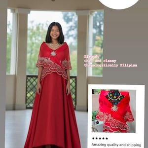 May include: A red Amparo Panuelo featuring intricate gold embroidery along the scalloped edges and a central decorative brooch. The garment is worn over a matching red floor-length dress with a V-neckline and a wide, draped shawl-style collar.