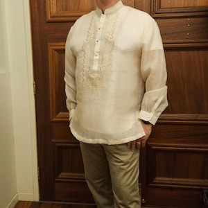 May include: A cream-colored, long-sleeved barong tagalog shirt featuring a mandarin collar, a button-down front placket, and intricate tonal embroidery along the chest and cuffs. The garment is made of a sheer, lightweight fabric.