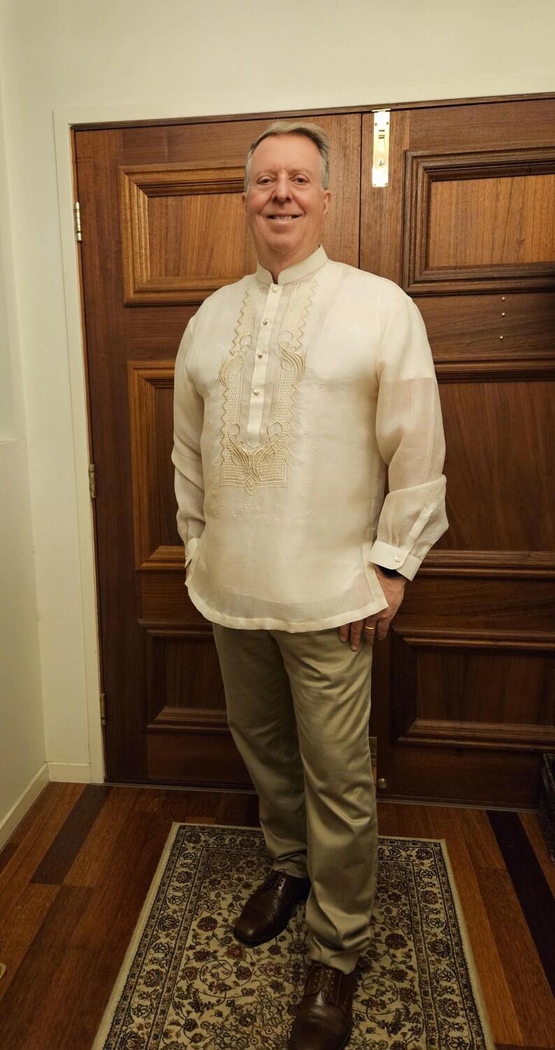 May include: A cream-colored, long-sleeved barong tagalog shirt featuring a mandarin collar, a button-down front placket, and intricate tonal embroidery along the chest and cuffs. The garment is made of a sheer, lightweight fabric.