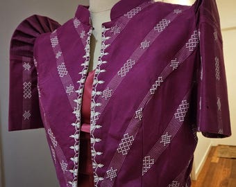 May include: A magenta jacket with white embroidered details and short sleeves. The jacket features a mandarin collar and diagonal striped patterns. The sleeves have a unique, structured design. The jacket is displayed on a mannequin.