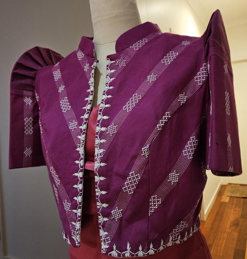 May include: A magenta jacket with white embroidered details and short sleeves. The jacket features a mandarin collar and diagonal striped patterns. The sleeves have a unique, structured design. The jacket is displayed on a mannequin.
