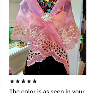 May include: A sheer pink organza shawl featuring intricate scalloped edges with floral embroidery and cutwork details. The wrap is secured at the centre with a cluster of white faux pearls and is draped over a black mannequin with a star pattern.