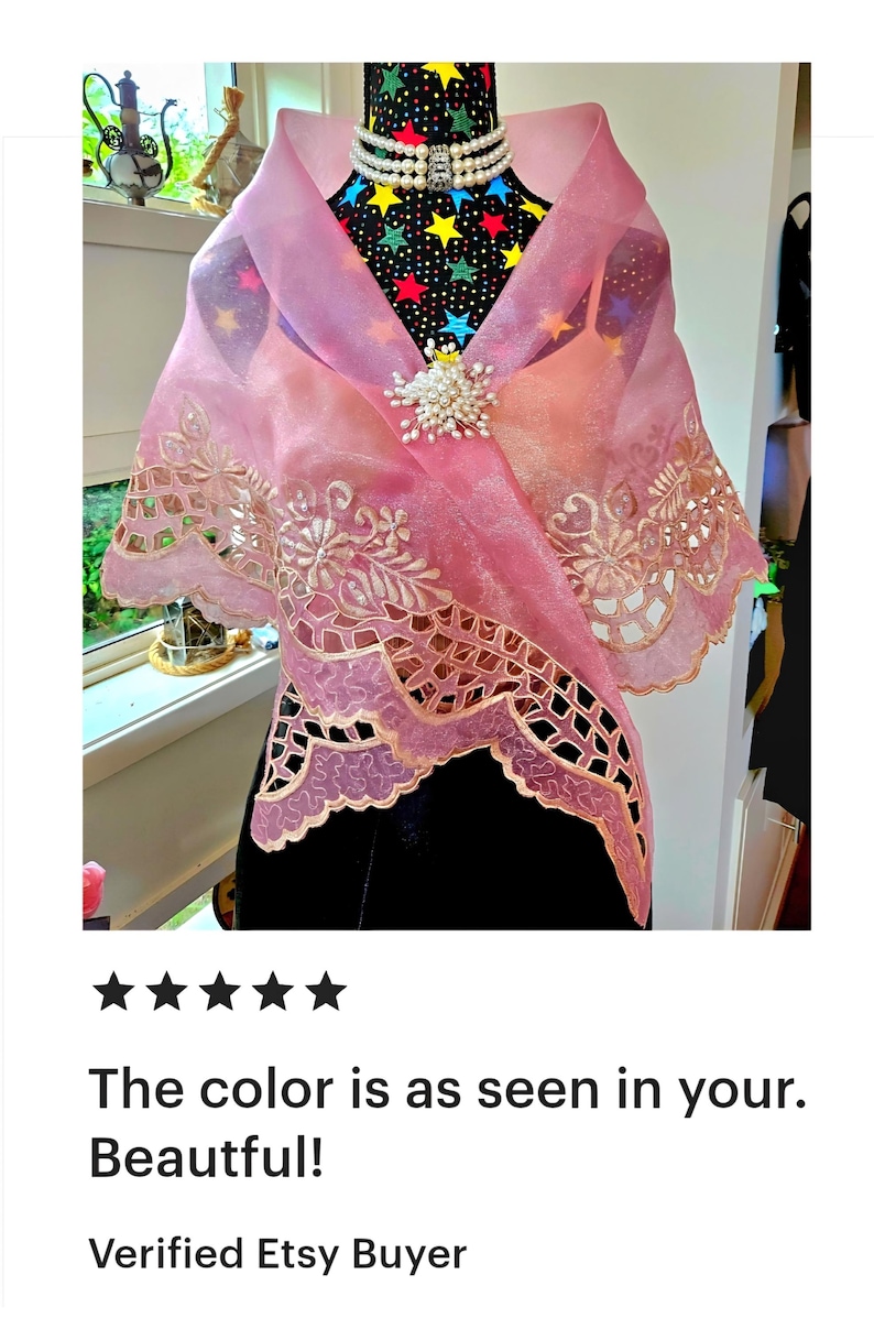 May include: A sheer pink organza shawl featuring intricate scalloped edges with floral embroidery and cutwork details. The wrap is secured at the centre with a cluster of white faux pearls and is draped over a black mannequin with a star pattern.