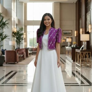 May include: A woman wearing a white dress and a purple bolero jacket with a white top underneath. The bolero has short sleeves and a patterned design. The background is a well-lit interior with marble flooring and large windows.