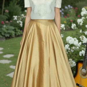 May include: A woman wearing a cream-colored top with embroidered details and a gold skirt. The skirt is floor-length and has a flowing, satin-like appearance. The background includes a garden with white flowers and a guitar.