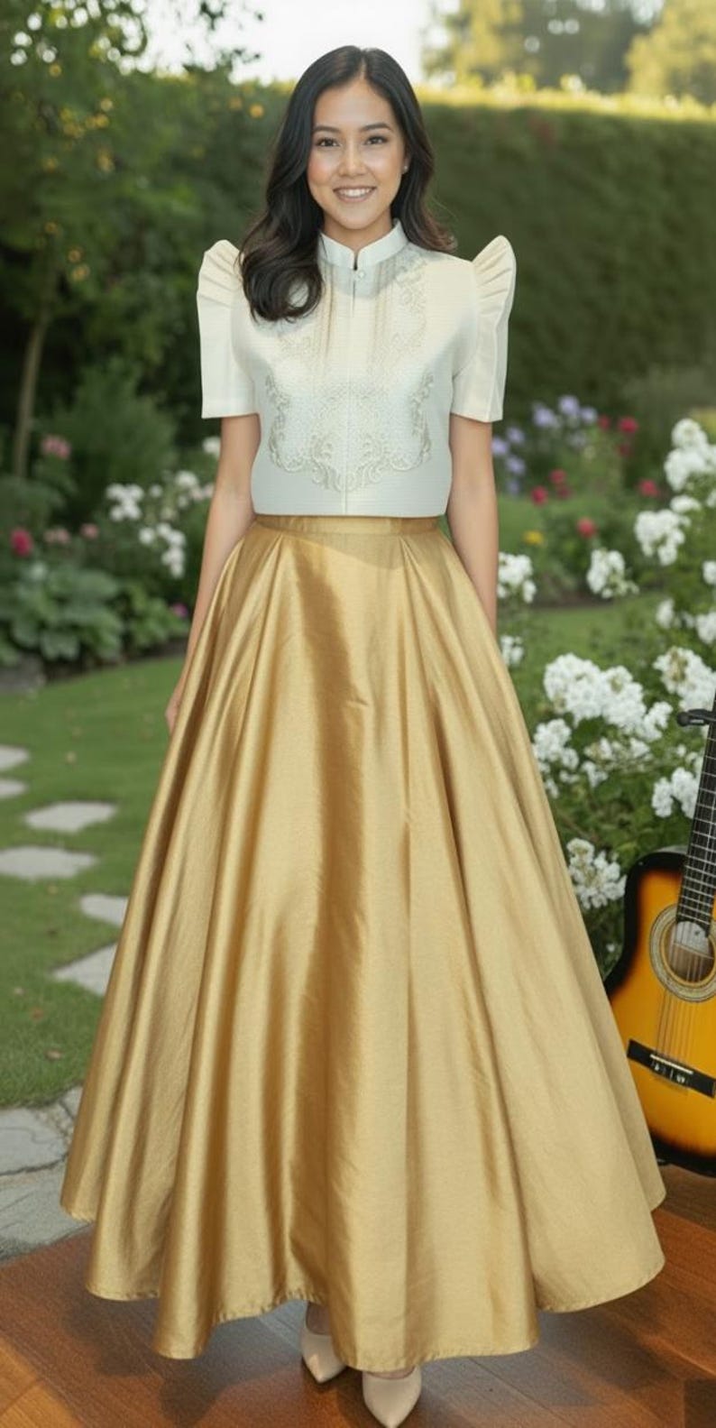 May include: A woman wearing a cream-colored top with embroidered details and a gold skirt. The skirt is floor-length and has a flowing, satin-like appearance. The background includes a garden with white flowers and a guitar.