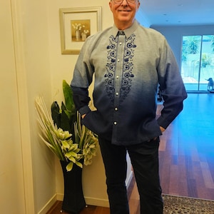 May include: A long-sleeved button-down shirt featuring a blue ombre gradient that transitions from light blue at the shoulders to dark blue at the hem. The shirt includes a dark blue floral embroidery pattern along the front placket and dark trousers.