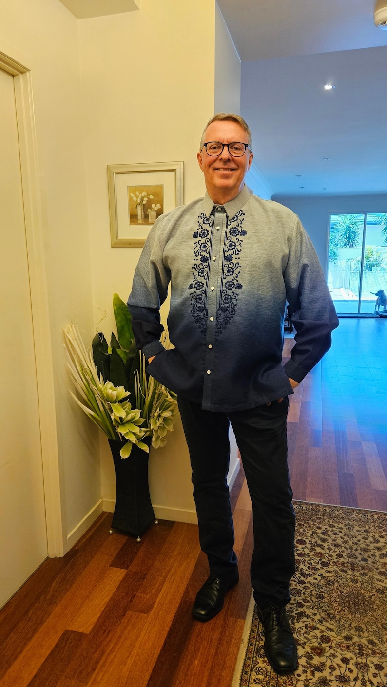 May include: A long-sleeved button-down shirt featuring a blue ombre gradient that transitions from light blue at the shoulders to dark blue at the hem. The shirt includes a dark blue floral embroidery pattern along the front placket and dark trousers.