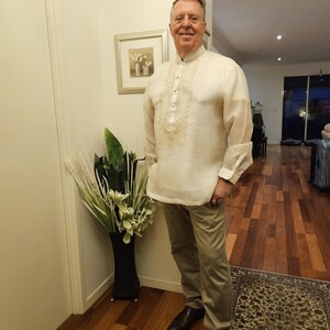 May include: A long-sleeved, off-white barong tagalog shirt featuring intricate, light-colored embroidery along the front placket and chest, paired with solid khaki-colored trousers and dark brown leather dress shoes.
