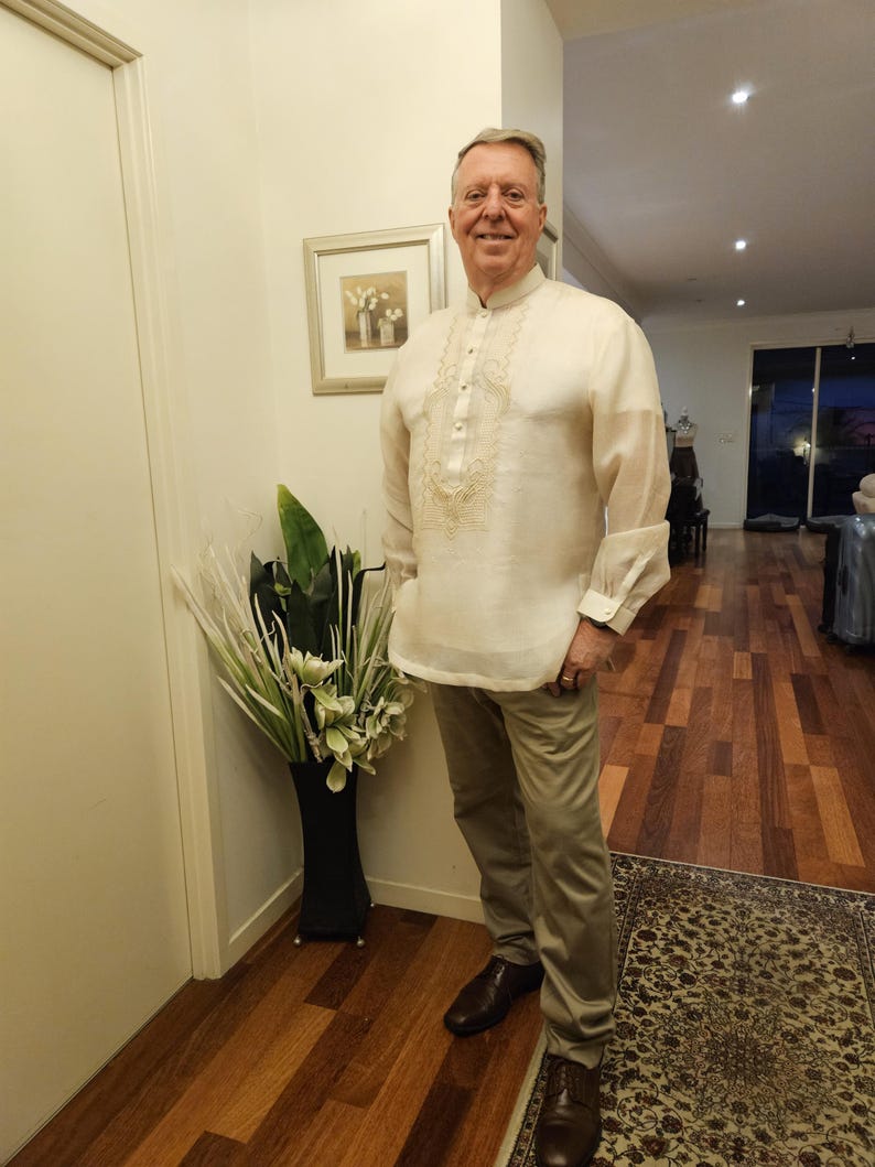 May include: A long-sleeved, off-white barong tagalog shirt featuring intricate, light-colored embroidery along the front placket and chest, paired with solid khaki-colored trousers and dark brown leather dress shoes.