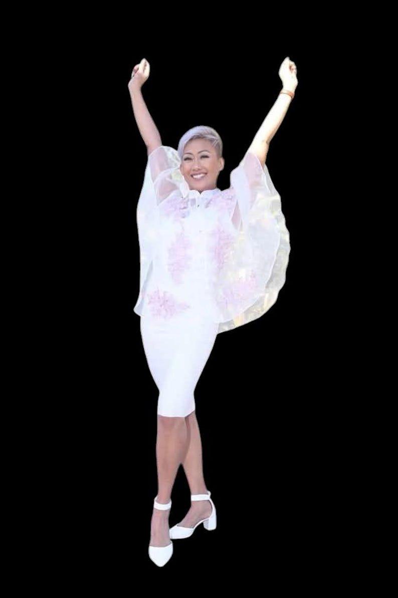 May include: A woman in a white dress with a sheer, floral-embroidered overlay, arms raised in a celebratory pose. She wears white heeled sandals. The background is black.