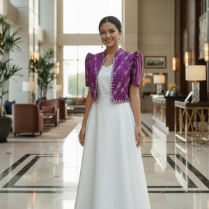 May include: A woman wearing a white floor-length dress and a purple bolero jacket with short sleeves. The jacket has a geometric pattern and a white trim. The woman is smiling in a well-lit, elegant interior.