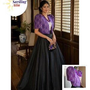 May include: A purple lace bolero jacket with structured puff sleeves worn over a white top and a long, voluminous black skirt. The outfit is accessorized with a purple beaded necklace and a small black clutch featuring a purple floral embroidery.