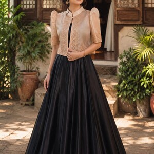 May include: A two-piece ensemble consisting of a black floor-length skirt and a cropped, beige, short-sleeved jacket. The jacket features a high collar, structured puff sleeves, and intricate embroidery along the front panels and sleeve hems.