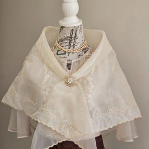 以下が含まれることがあります： スカラップ状の縁に繊細な花柄の刺繍が施された、オフホワイトの透け感のあるオーガンジーショール。中央は小さな白いパールと透明なクリスタルで飾られた丸いブローチで留められており、パールネックレスの上に着用されています。