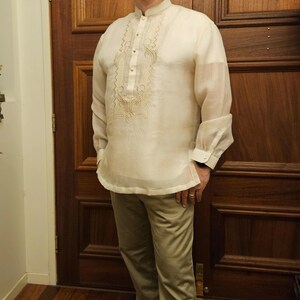 May include: A cream-colored, long-sleeved barong tagalog shirt featuring a mandarin collar, a button-down front placket, and intricate tonal embroidery on the chest, paired with light tan trousers and dark brown leather dress shoes.