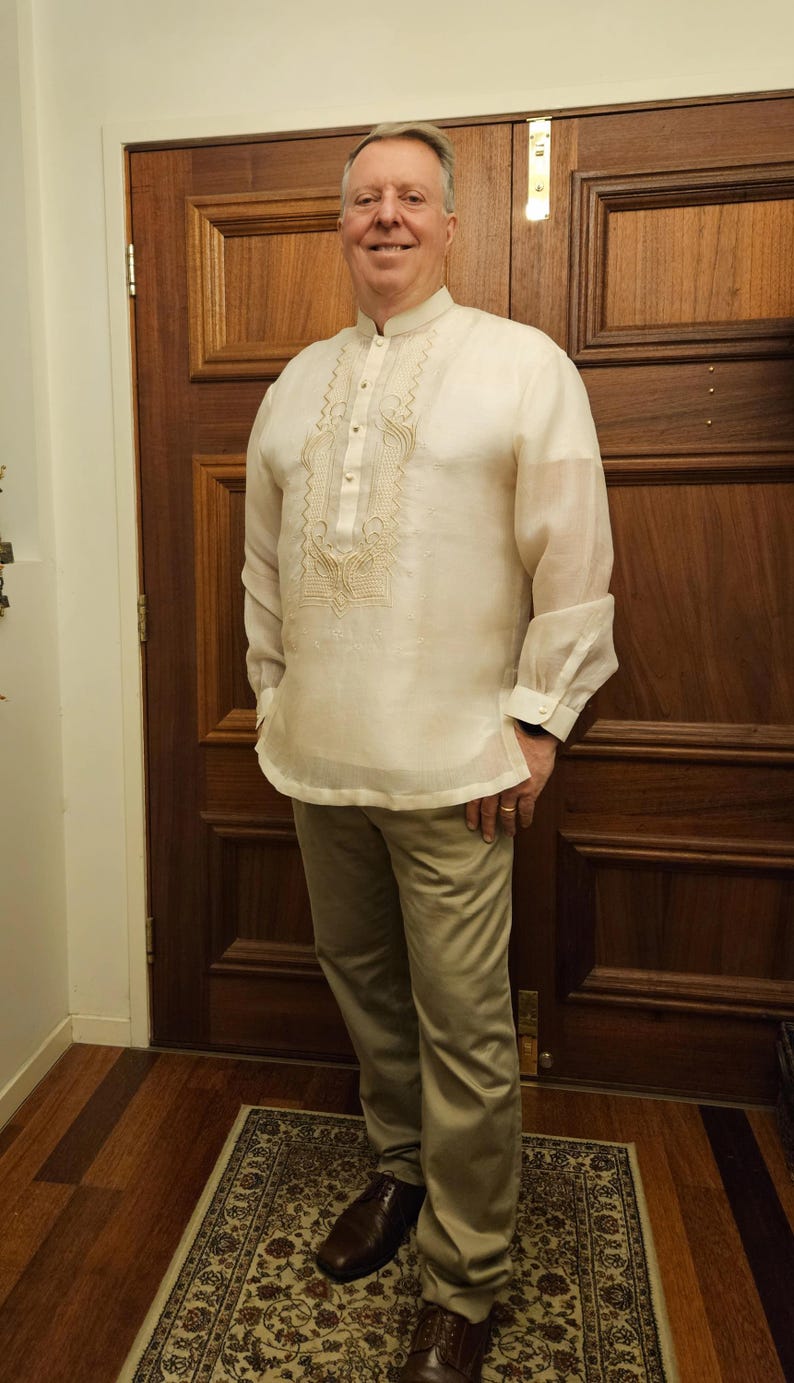May include: A cream-colored, long-sleeved barong tagalog shirt featuring a mandarin collar, a button-down front placket, and intricate tonal embroidery on the chest, paired with light tan trousers and dark brown leather dress shoes.